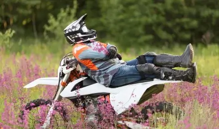 A dirt biker relaxes on their bike in a field of purple flowers.