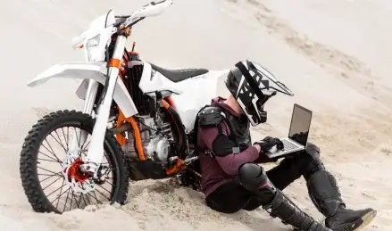 A dirt biker works on a laptop while sitting in sand beside their bike.